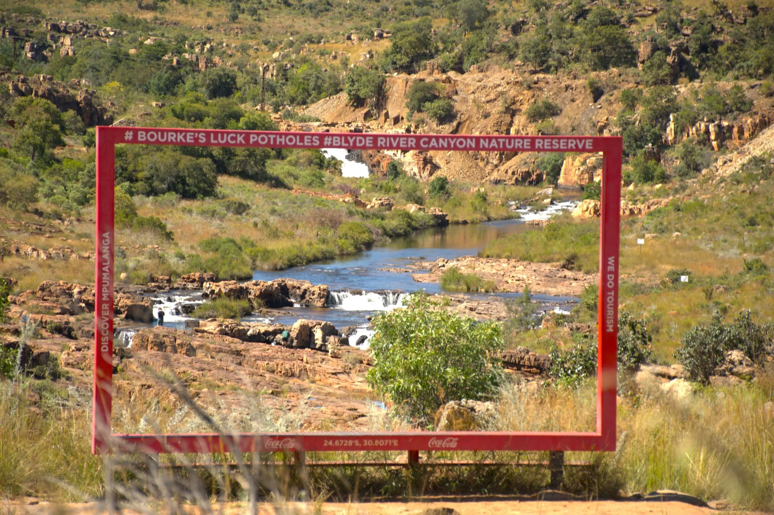 Looking through a bold red picture frame viewfinder at Bourke's Luck Potholes in the Blyde River Canyon Nature Reserve, Mpumalanga — the frame bears the words Blyde River Canyon and frames the canyon and rolling grassland landscape beyond