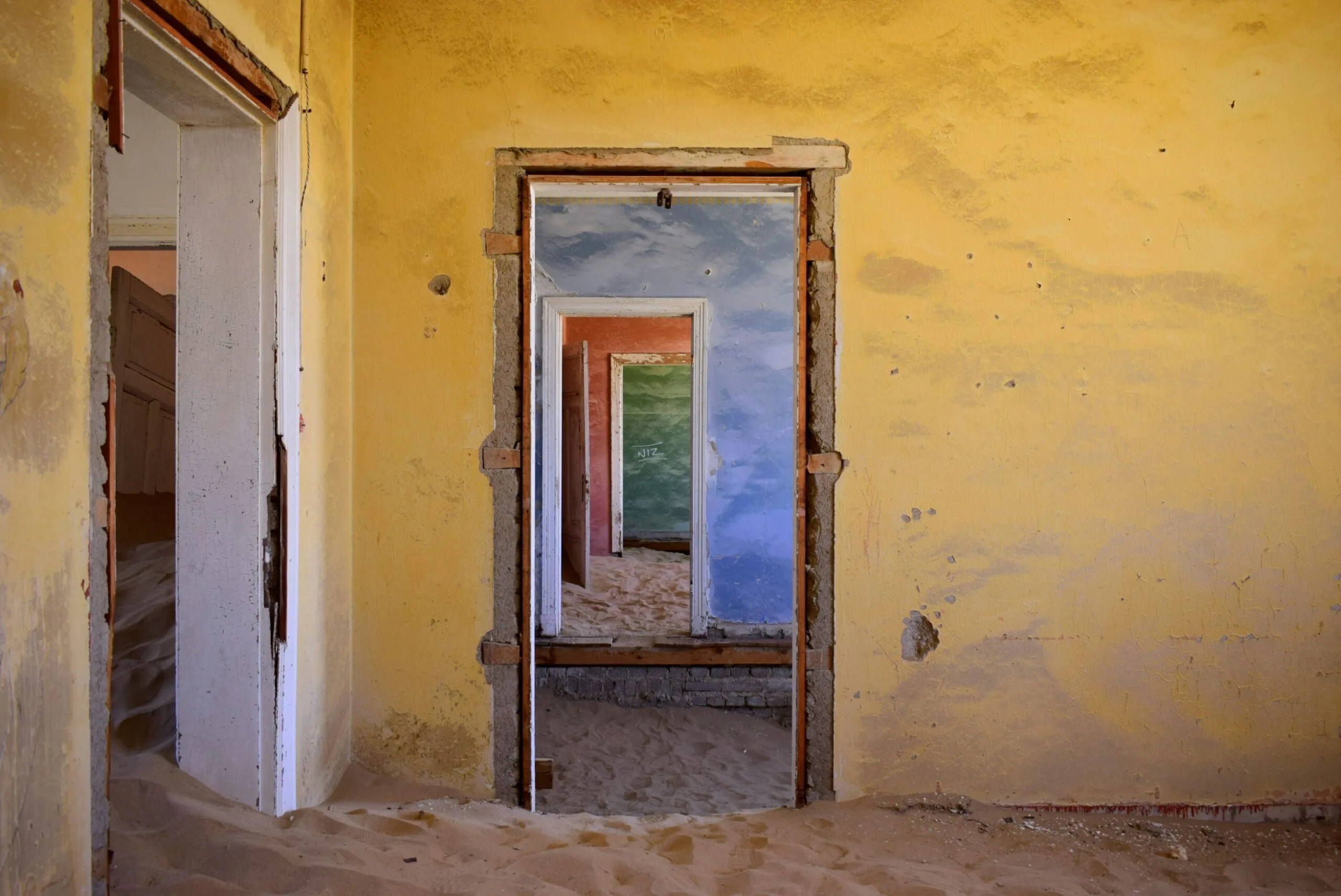 Looking down a sand-invaded Kolmanskop corridor: yellow front room, blue middle room, red back room, and a green cross-corridor visible through doorless openings