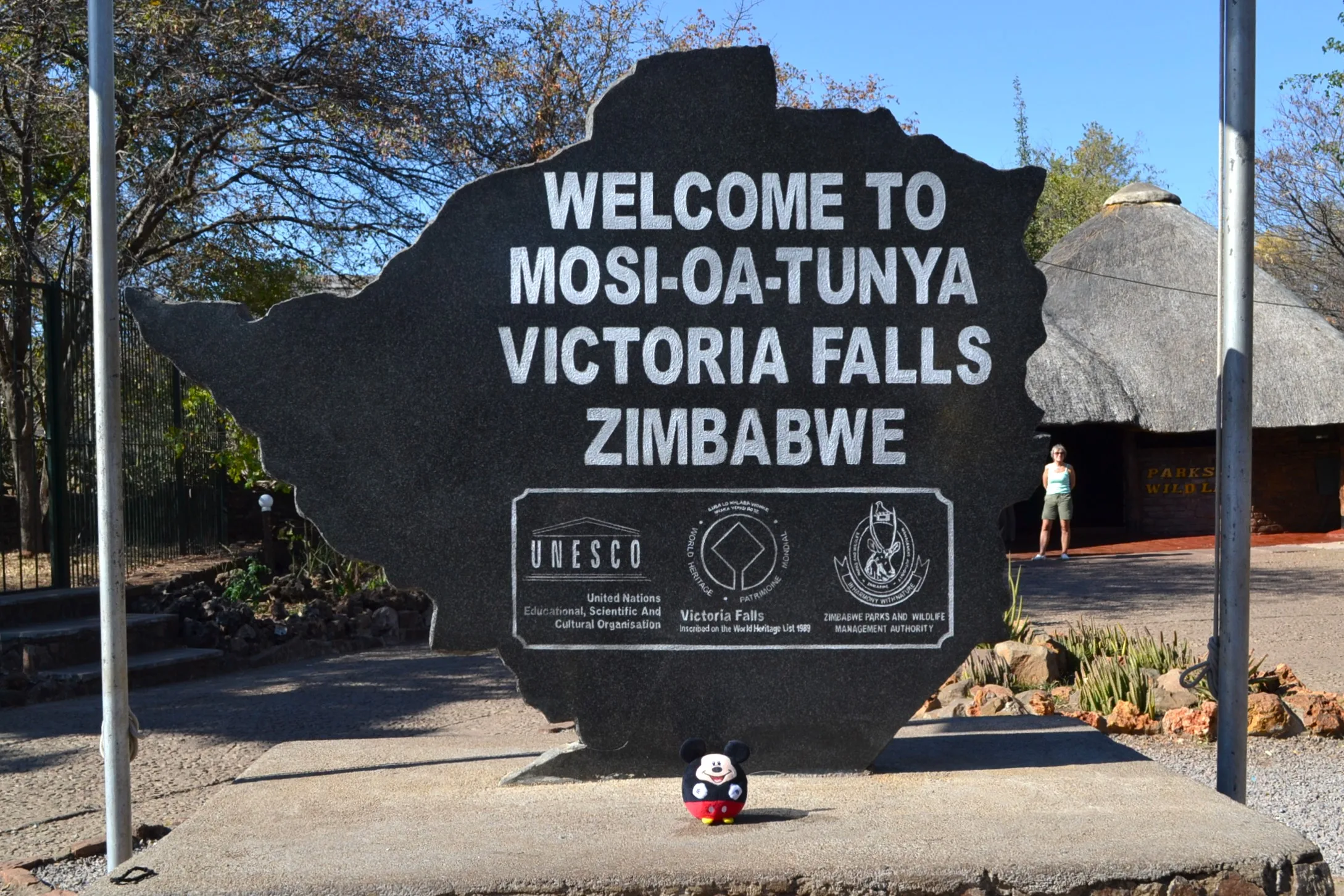 Flat granite rock sculpture shaped like the silhouette of Zimbabwe, bearing the inscription WELCOME TO MOSI-OA-TUNYA VICTORIA FALLS ZIMBABWE, with logos for UNESCO, Victoria Falls, and Zimbabwe Parks and Wildlife; a visitor stands in the background beneath a thatched shelter