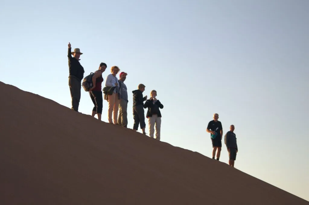 Two tourists waving from the crest of a dune as the sun sets behind them over the Namib